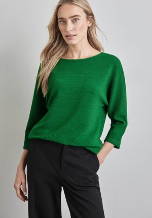 Groene gestructureerde top met een brede halslijn en driekwartmouwen, gecombineerd met zwarte broek. De stof heeft horizontale ribbels.