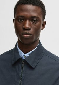 Giacca zip-blu navy con colletto a punta sopra una camicia azzurro chiaro con bottoni. Tessuto liscio, design minimalista e nessun motivo visibile.