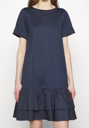 Kvinde iført en knælængde marineblå kjole med korte opsmøgede ærmer og en dobbeltlags flæsekant.