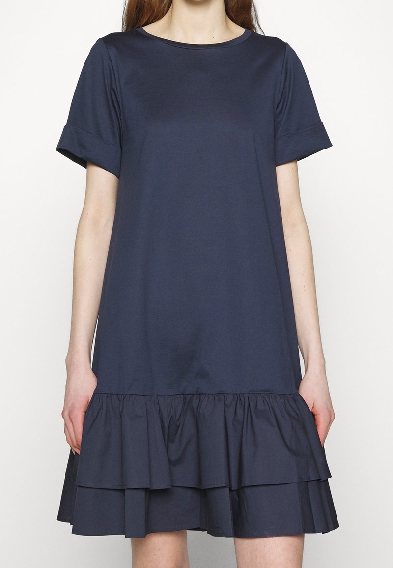 Femme portant une robe bleu marine longueur genou avec des manches courtes retroussées et un ourlet à volants à double couche.