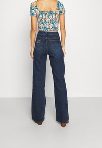 Blue denim high-waisted jeans with a wide-leg cut, featuring two back pockets, a logo tag, and a smooth texture.