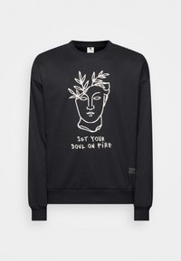 Sudadera negra de manga larga, con un diseño de arte lineal blanco de un rostro adornado con hojas y el texto "PON TU ALMA EN LLAMAS."