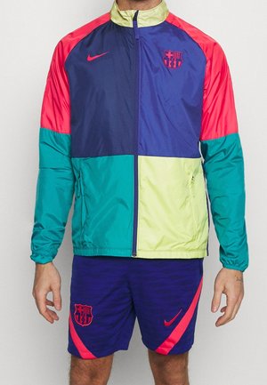 Mann trägt eine mehrfarbige (marineblau, rot, türkis, gelb) FC Barcelona Jacke und marineblaue Shorts mit roten Nike-Logos und FC Barcelona Wappen.