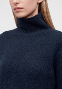 Gros plan d'une femme portant un pull à col roulé bleu foncé et duveteux avec de longs cheveux blonds lisses visibles.