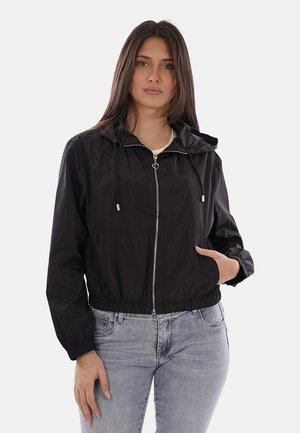 Frau trägt eine schwarze Kapuzenjacke mit Reißverschluss und Kordelzug sowie hellblaue Jeans, steht mit einer Hand in der Tasche vor einem schlichten Hintergrund.