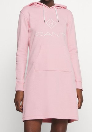 Femme portant une robe sweat à capuche rose clair avec poche avant et logo blanc "GANT" sur la poitrine.