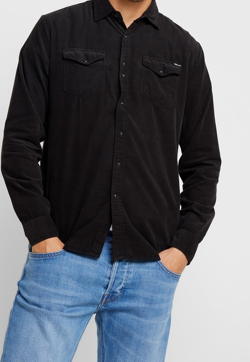 Camisa negra abotonada de tela suave con dos bolsillos en el pecho, mangas largas y corte recto, combinada con vaqueros de mezclilla azul claro.