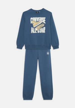 Conjunto de sudadera y pantalones de chándal azules con texto "Converse All Star" y un gráfico de zapatilla beige en la sudadera, y el logo en los pantalones.