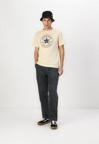 Camiseta beige con un logo de Converse en rojo y azul, sombrero de cubo negro, pantalones de pana oscuros y zapatillas negras con cordones blancos y suelas marrones.