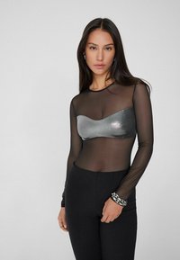 Top negro transparente de manga larga con un busto estilo bandeau metálico plateado. Combinado con pantalones negros ajustados. Pulsera de cadena plateada como acento.