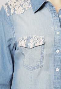 Chemise en denim bleu clair avec des accents en dentelle sur les épaules et les rabats de poche, dotée d'un col classique et d'une fermeture à boutons.