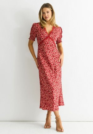 Woman in a red floral midi dress with puff sleeves and lace trim, standing against a white background wearing beige ankle-strap heels.
