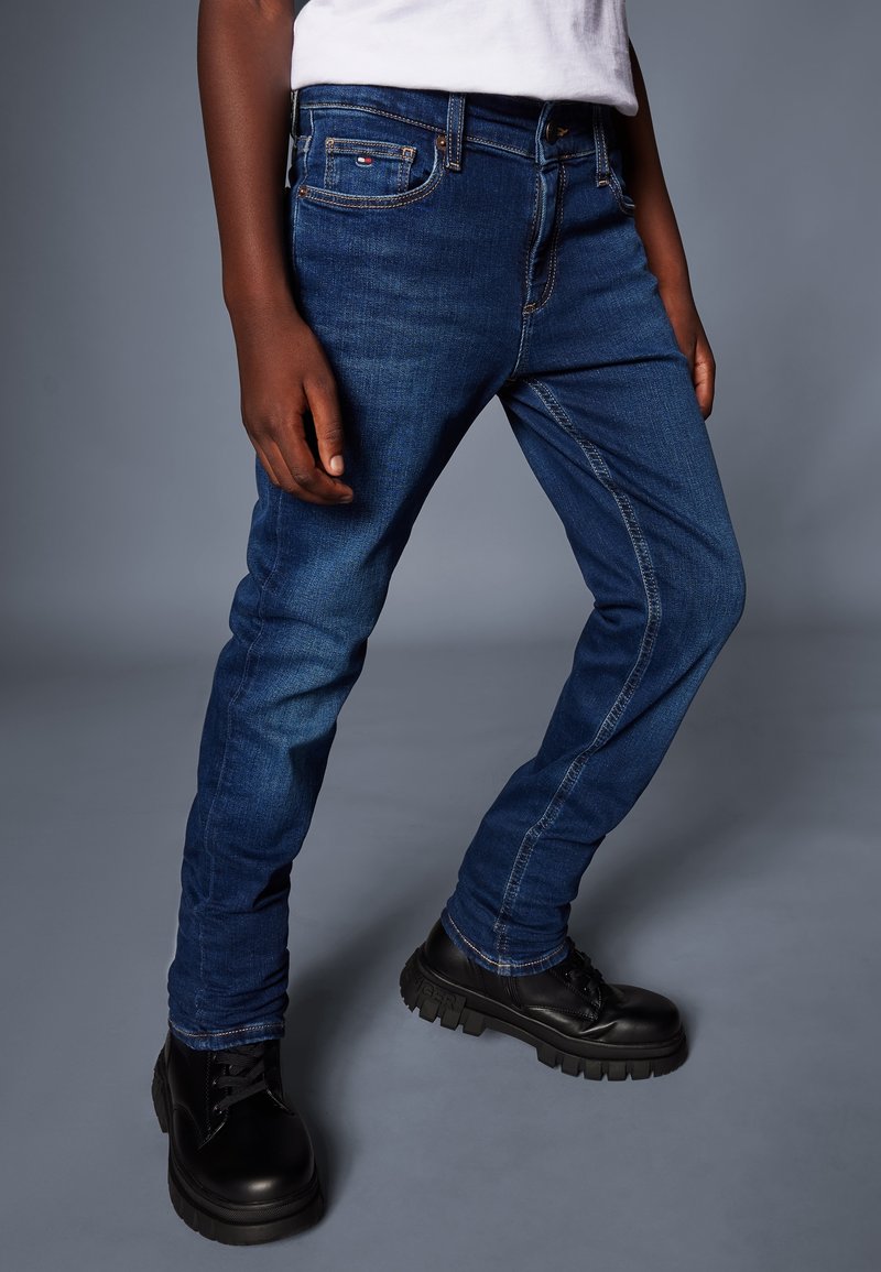 Personne portant un jean slim bleu foncé, des chaussures noires à lacets épaisses, et une chemise blanche, debout devant un fond gris.