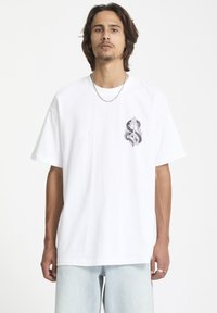 T-shirt en coton blanc avec un design graphique en violet et gris sur la poitrine gauche, à manches courtes et à coupe décontractée.
