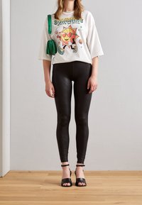 Vit oversized T-shirt med en färgglad solgrafik, matchad med svarta leggings i ormskinnsmönster och svarta öppna klackskor. Grön handväska som accent.