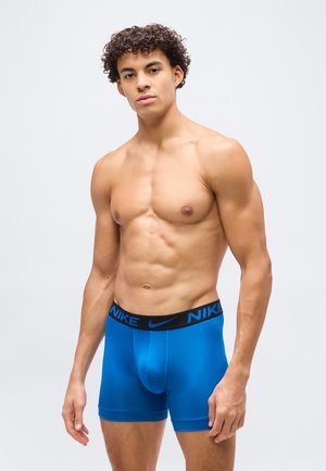 Γυμνός από τη μέση και πάνω νεαρός άνδρας με σγουρά μαλλιά που φοράει μπλε εσώρουχα μπόξερ της Nike, στέκεται μπροστά σε ένα απλό ανοιχτό γκρι φόντο.