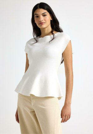 Kvinde med langt mørkt hår iført en tætsiddende hvid ærmeløs top med peplumkant og beige bukser mod en ensfarvet baggrund.