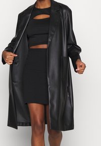 Cappotto lungo in faux pelle nero con una sola tasca, indossato sopra un crop top a coste nero e una gonna coordinata. Texture liscia e lucida.
