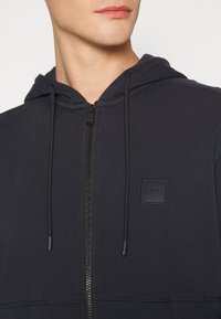 Sudadera con cremallera de color azul marino, con capucha, cordones ajustables, puños de canalé, costuras planas y un pequeño parche de logo en el pecho.