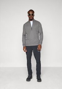 Felpa grigia con zip, pantaloni leggeri neri con tasche laterali e stivali neri. Il modello indossa occhiali da sole e ha i capelli intrecciati.