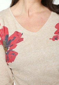 Beżowy sweter z dekoltem w serek i kwiatowym haftem w kolorze czerwonym i granatowym. Posiada teksturowany materiał z kwiatowymi akcentami i małymi złotymi detalami.