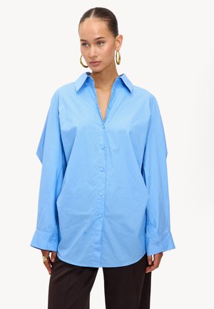 Camisa oversize azul claro de algodón, con cierre de botones en la parte delantera, cuello afilado y hombreras caídas con puños anchos.