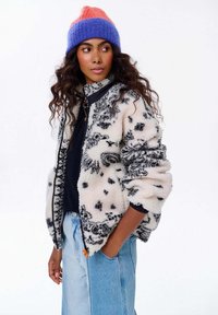 Chlpatá, čierno-bielo vzorovaná fleece bunda so zipsom vpredu a stojatým golierom; spojená s námorníckou košeľou a svetlomodrými džínami.