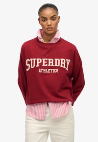 Sudadera marrón con "SUPERDRY ATHLETICS" en texto color crema. Debajo, se asoma una camiseta a rayas rosas y blancas. Pantalones crema neutros.