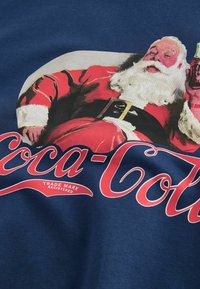 T-shirt en coton bleu marine présentant un graphique d'un Santa souriant dans un costume rouge avec des bordures blanches, tenant une bouteille de Coca-Cola sous le logo.