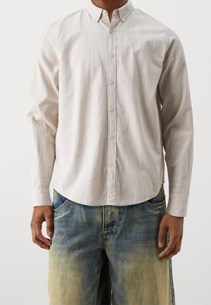 Mann trägt ein weißes, langärmeliges, strukturiertes Hemd mit Knöpfen und ausgewaschene blaue Jeans, steht vor einem einfarbigen Hintergrund.