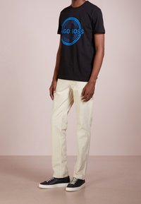 T-shirt en coton noir avec un logo "HUGO BOSS" bleu, associé à un pantalon beige clair et des baskets noires avec semelles et lacets blancs.