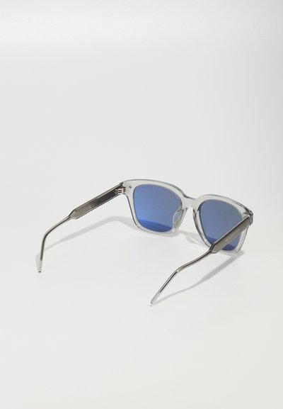 Ochelari de soare din plastic transparent cu lentile albastre și brațe din metal. Formă pătrată cu accente texturate pe tâmple. Design curat, modern.
