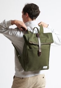 Mochila de lona verde con diseño de parte superior enrollable, correa de cuero marrón, cremallera dorada y bolsillo frontal. Correas ajustables y asas reforzadas.