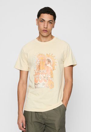 Jeune homme aux cheveux courts et bouclés porte un tee-shirt graphique beige avec un soleil, des étoiles et le texte « Here to Love », assorti à un pantalon cargo olive.