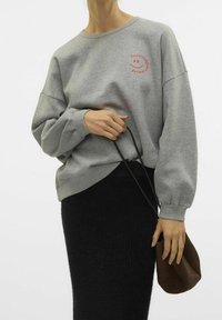 Sudadera gris con cuello redondo y corte holgado, que presenta texto rojo y un gráfico de una carita sonriente. Combinada con una falda negra texturizada y un bolso marrón.