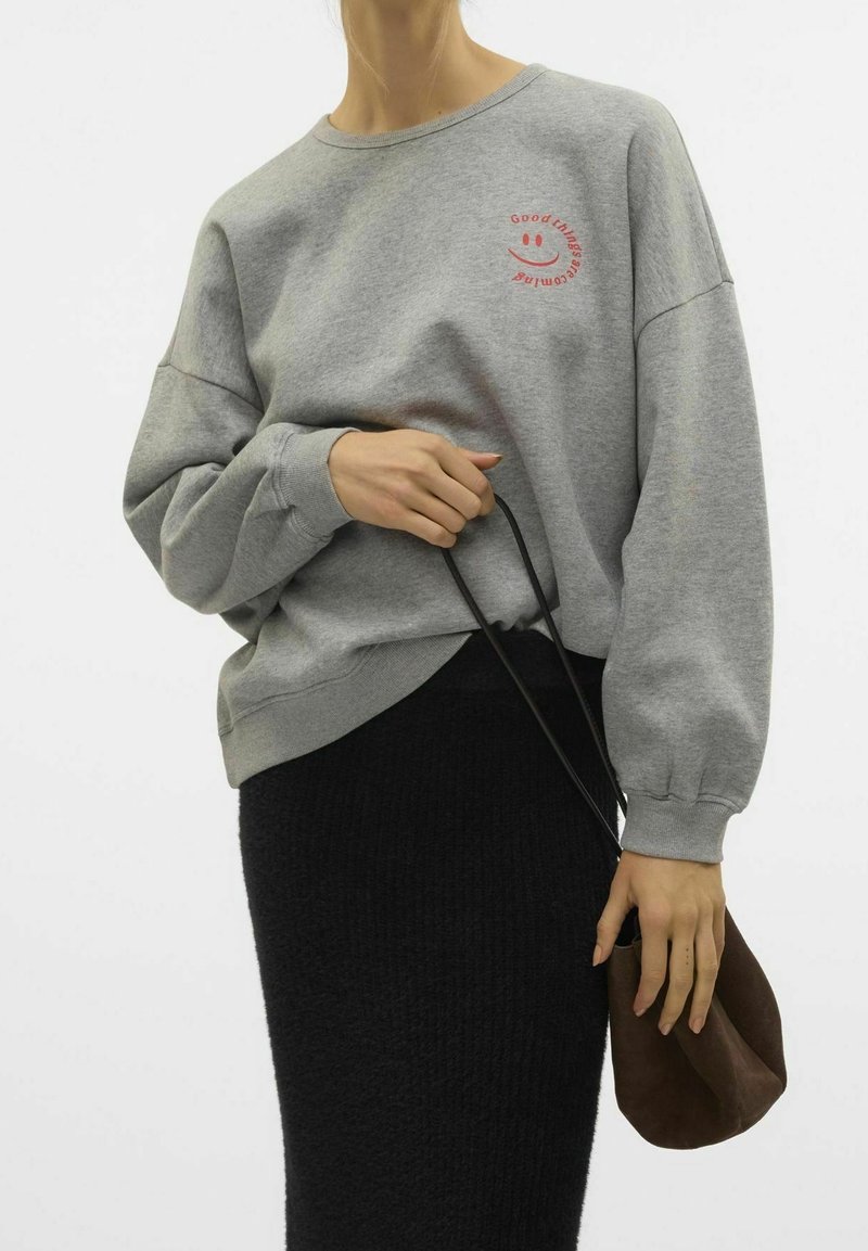Sudadera gris con cuello redondo y corte holgado, que presenta texto rojo y un gráfico de una carita sonriente. Combinada con una falda negra texturizada y un bolso marrón.