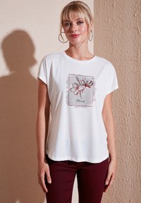 Witte t-shirt met korte mouwen, voorzien van een bloemendessin in rood en grijs. Zacht materiaal met een relaxte pasvorm en ronde halslijn.