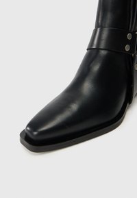 Botín de cuero negro hasta el tobillo con punta afilada, que presenta una correa y detalles en metal plateado. Textura suave y color negro sólido.