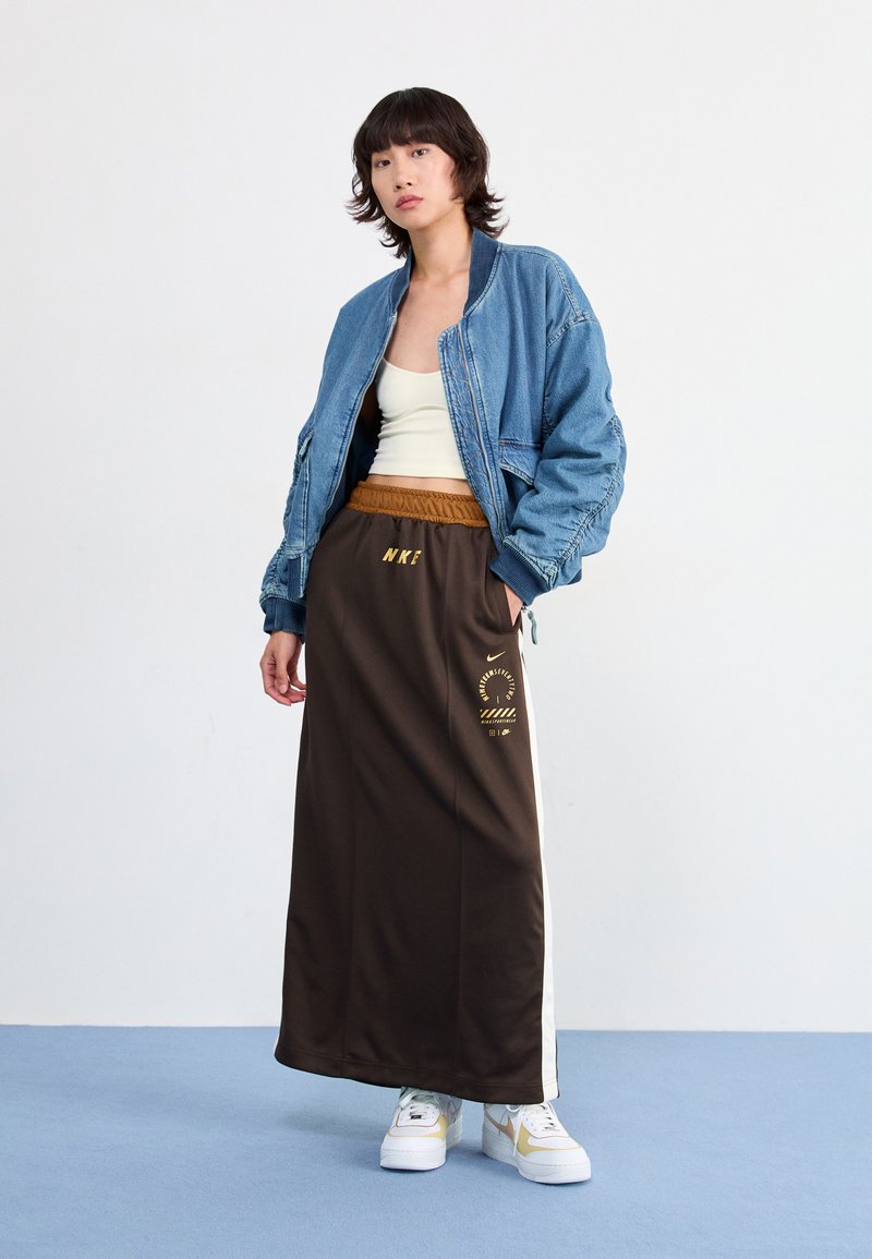 Chaqueta bomber de denim, top de tirantes crema y falda maxi marrón con estampado dorado. Zapatillas blancas con acentos dorados. Texturas suaves y ajuste relajado.