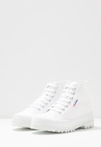 Sneakers alte bianche in tela con suola in gomma testurizzata, caratterizzate da occhielli in metallo e un piccolo logo blu e rosso sul lato.