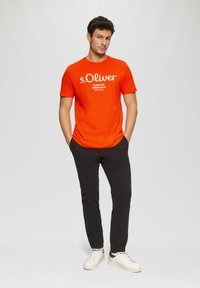 Jasně oranžové bavlněné tričko s bílým logem "s.Oliver" a textem "ELEVATED ESSENTIALS". Spárováno s černými kalhotami a bílými teniskami.