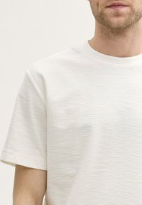 Fehér, kerek nyakú póló, texturált anyagból készült; rövid ujjakkal és laza fazonnal rendelkezik, finom, egyenetlen felületi mintázattal.