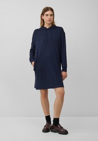 Frau steht in einem navyblauen Hoodie-Kleid mit Taschen, schwarzen Socken und dunkelburgunderfarbenen Slippern vor einem einfarbigen grauen Hintergrund.