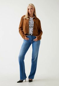 Brun cropped jakke i ruskind, grafisk t-shirt, lyseblå straight-leg jeans og sorte hæle. Glat tekstur, knaplukning på jakken.