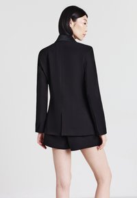 Donna con capelli neri corti che indossa un blazer nero e pantaloni corti coordinati, in piedi contro uno sfondo grigio chiaro semplice, rivolta di lato.