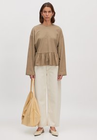 Beige Baumwoll-Peplum-Oberteil mit langen Ärmeln und rundem Halsausschnitt, kombiniert mit einer weiten cremefarbenen Hose und einer weichen gelben Tasche. Kontraste in den Texturen.