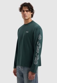 Donkergroene katoen T-shirt met lange mouwen, voorzien van een klein logo op de borst en vetgedrukte "Pure Path" tekst die langs de linkermouw is gedrukt.