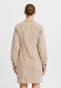 Beige kordflöjdsklänning med en skräddarsydd silhuett, långa ärmar, knappade manschetter och en sömdetalj vid midjan. Bakåtvyn visas.