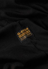Zwarte stof met een gestructureerde afwerking met een gouden geborduurd label waarop "G-Star Originals RAW Denim" staat.