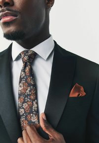 Blomstermønstret slips i brun, beige og marineblå, parret med en hvid skjorte og sort jakkesæt. En orange lommetørklæde er synlig.